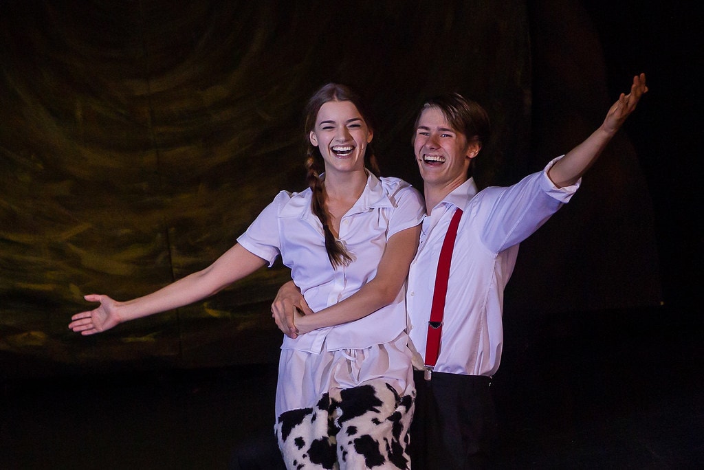 Gypsy — Louise in her cow-print pajamas and the young man in the white shirt and red suspenders stand arm in arm with arms spread wide and broad smiles before the golden hay bale backdrop in a triumphant pose