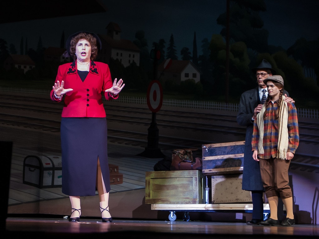 Gypsy — Rose in her red blazer gestures with both hands raised toward the audience before the painted train station backdrop as the man in the grey overcoat stands with his arm around the young woman in the plaid jacket at right
