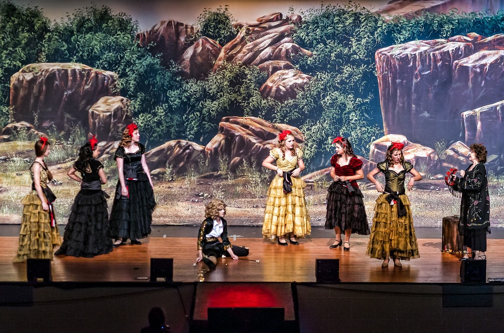 Gypsy — eight women in a Spanish dance number: six in black-and-gold ruffled flamenco skirts with red rose hair pieces stand in two lines, while a woman in a gold dress and a woman in a deep red velvet gown face each other at center, and Rose in a black embroidered shawl holds a tambourine at far right before a sweeping painted rocky landscape backdrop