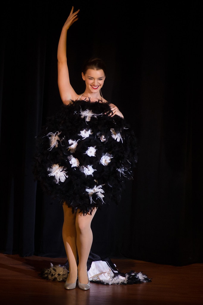 Gypsy — Louise in her showstopper black feather costume: a voluminous strapless ball of black feathers and white flower accents, one arm raised high with a broad smile as the white feather train pools at her feet on the bare stage