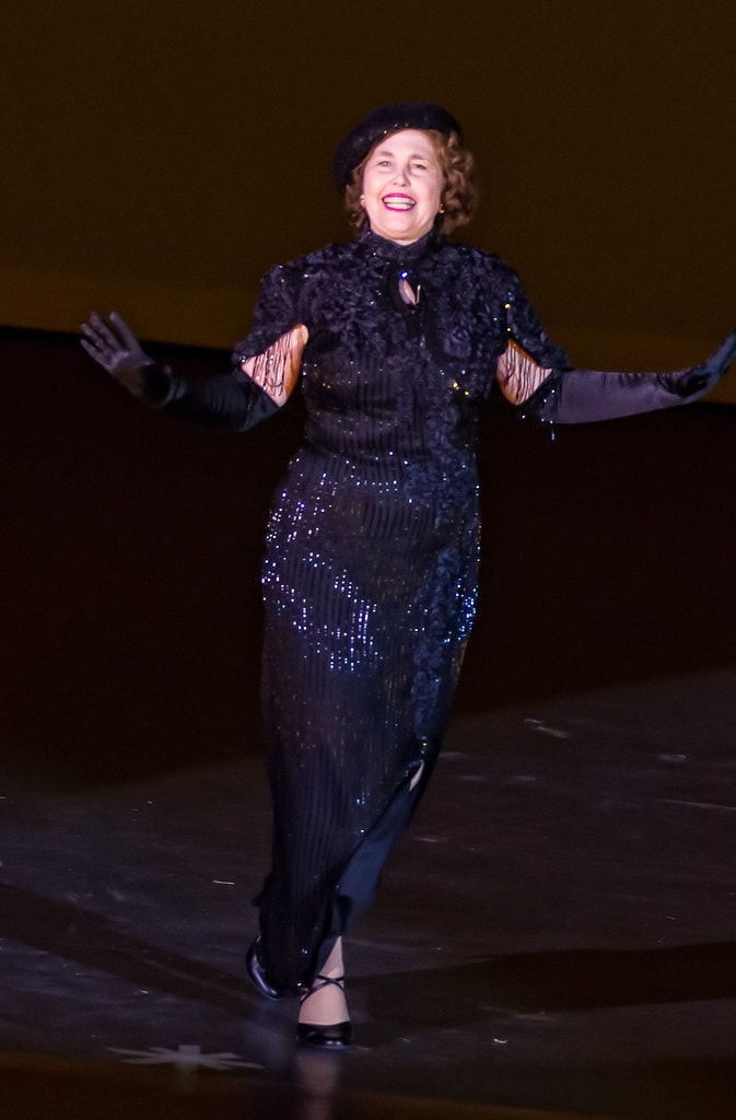 Gypsy — Rose's final moment: Mama Rose performs alone on a bare amber-lit stage in a full-length black beaded gown with a black fur-trimmed jacket and long black gloves, arms spread wide with a radiant smile, the spotlight catching the blue sequin details on her dress