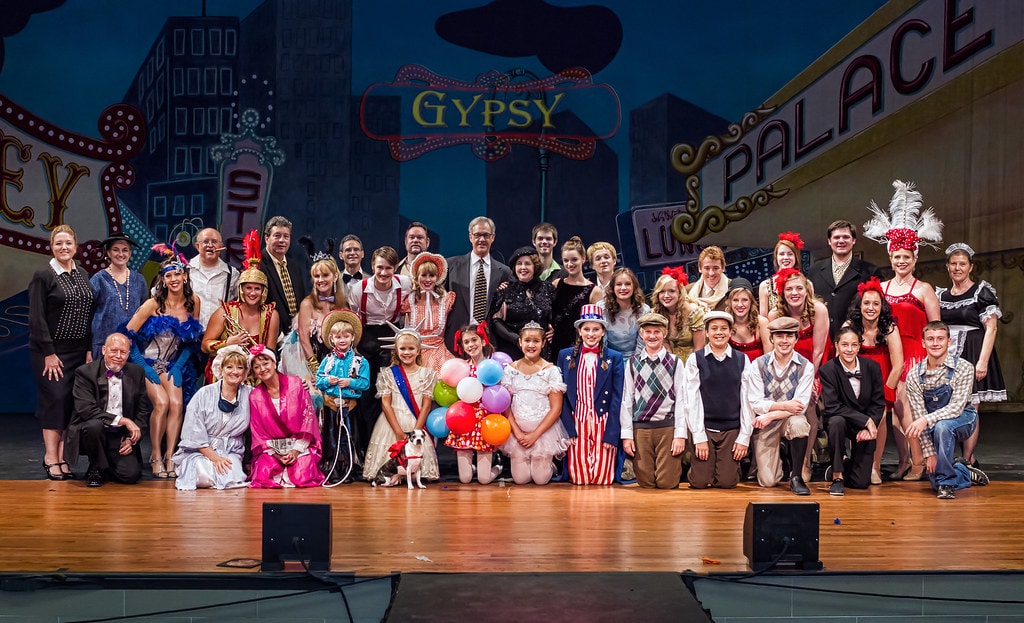 Gypsy — the full company photo: the entire cast of over 40 performers poses on stage before the Gypsy Palace backdrop with the show title in neon lights, from the children in their vaudeville costumes in the front row to the adult principals and ensemble in full costume behind them, a small white dog with a balloon visible at center front