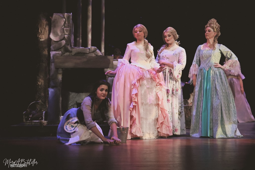 Into the Woods — Cinderella kneels by the fireplace as her stepsisters and stepmother stand over her in the cottage set