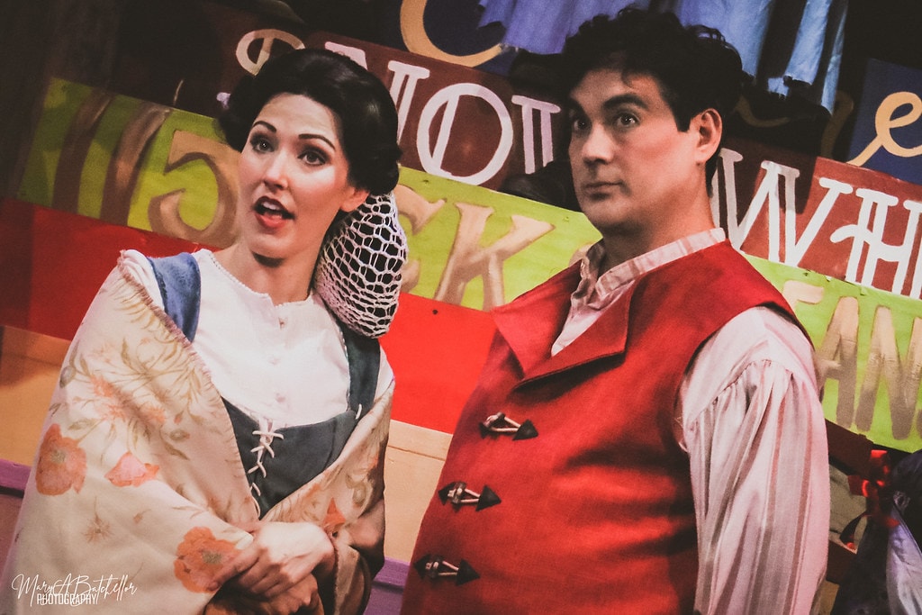 Into the Woods — the Baker's Wife and the Baker stand together before the colorful storybook backdrop