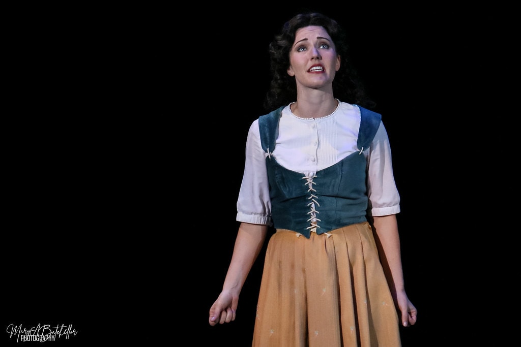 Into the Woods — the Baker's Wife in a green vest and tan skirt sings a close-up solo against a black backdrop