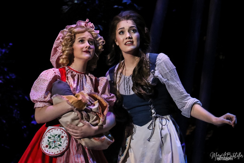 Into the Woods — Little Red Riding Hood and Cinderella stand together looking upward in the dark enchanted forest