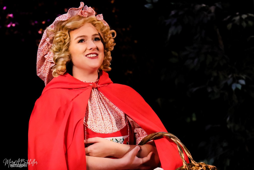 Into the Woods — Little Red Riding Hood in her red cape smiles warmly while holding her wicker basket in the dark forest