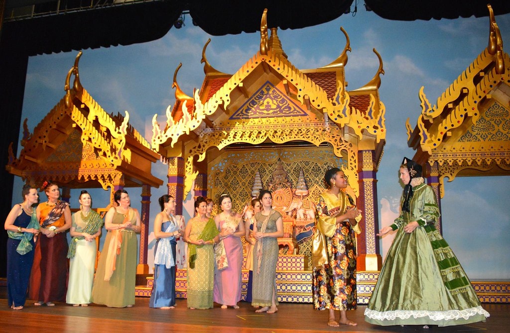 The King & I — Anna in a green Victorian gown faces a line of the King's wives in colorful Thai silk dresses before the ornate golden temple set with blue sky backdrop