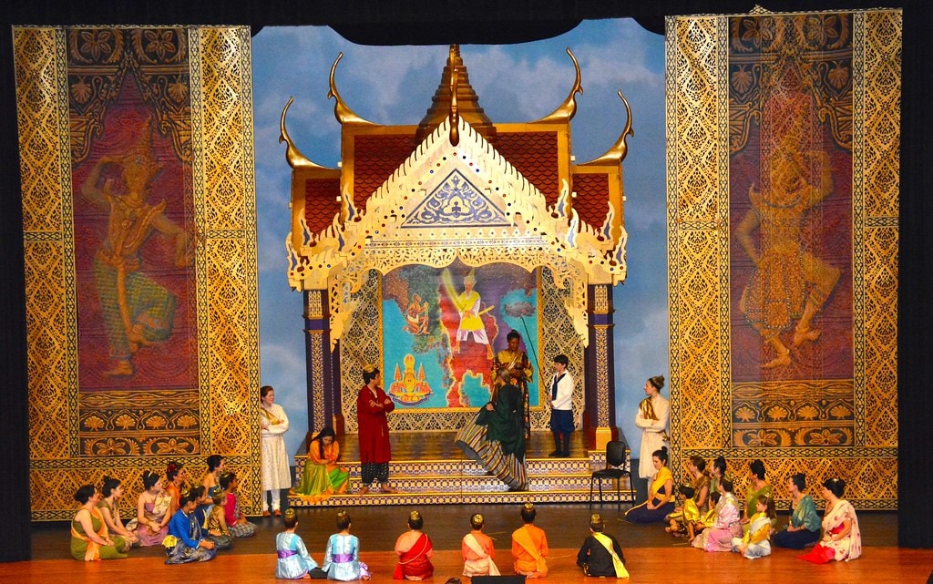 The King & I — the full ensemble of wives and children seated on the floor before the golden temple set as four standing figures including the King and Anna converse at center stage