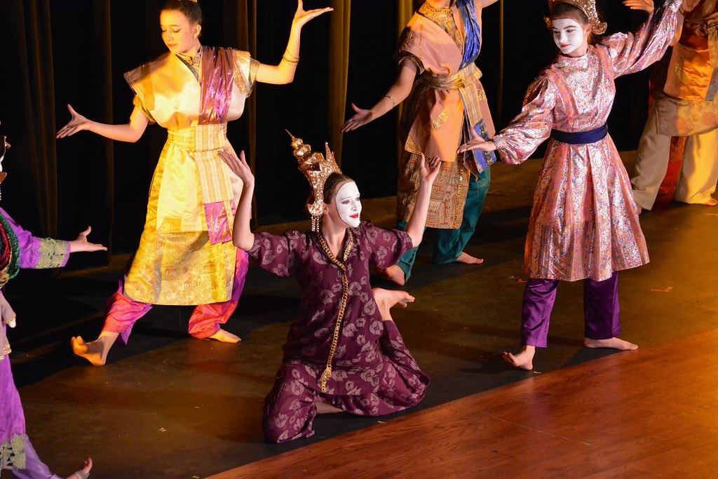 The King & I — a performer in a white-faced mask and purple floral robe kneels with arms raised as the full ensemble in Thai-inspired costumes dances around her in the Small House of Uncle Thomas ballet
