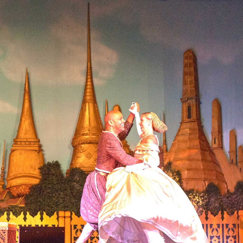 The King & I — the King in a burgundy Siamese costume lifts Anna in her golden ball gown as they dance together before the painted Bangkok temple spires backdrop