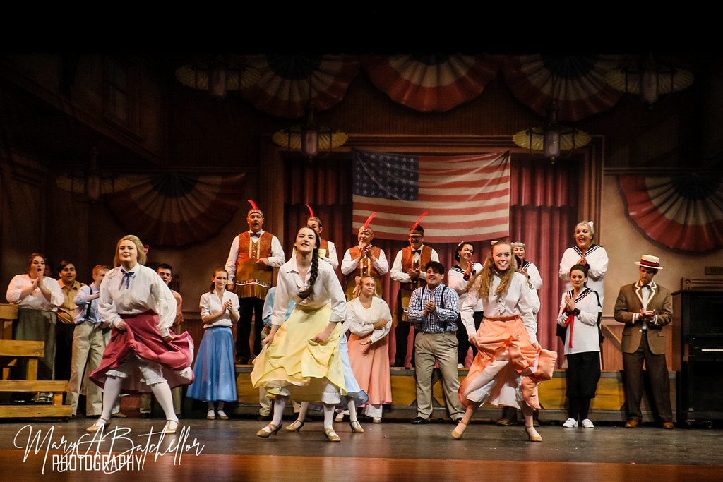The Music Man — two young women in colorful skirts leap as the full ensemble cheers