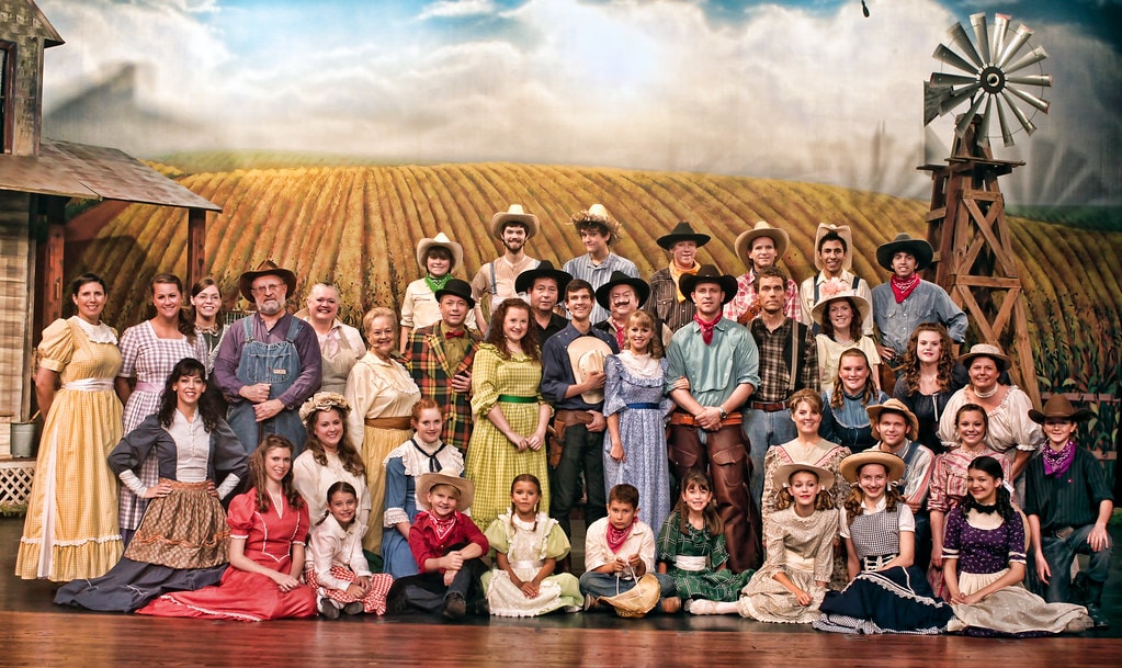 Oklahoma! — the full company photo: over 30 cast members in period prairie costumes pose on stage before the golden wheat field and windmill backdrop, from the children seated on the floor to the adult ensemble standing in rows behind them