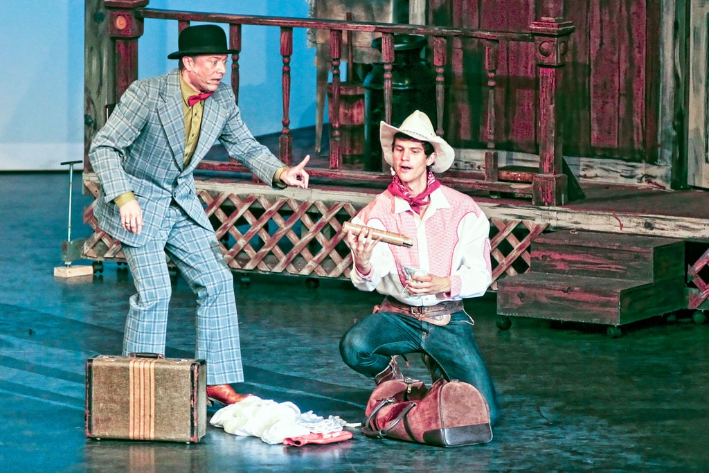 Oklahoma! — Ali Hakim in his grey plaid suit and black bowler hat points accusingly at Will Parker kneeling on the floor examining a rifle, Will's duffel bag and scattered belongings around him before the weathered farmhouse porch set