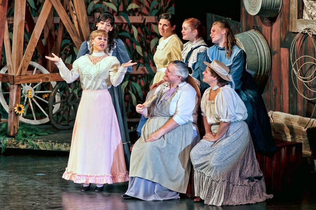 Oklahoma! — Laurey in a white blouse and pink skirt stands center with arms spread wide singing to six women in prairie dresses gathered around her in the barn set, a wagon wheel and sunflowers visible at left