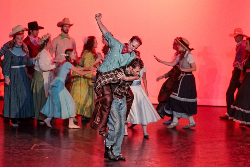 Oklahoma! — the barn dance brawl: Curly in his teal shirt lifts Jud Fry in his plaid shirt off the ground in a bear hug as the full ensemble of cowboys and women in prairie dresses swirl around them in the pink-lit dance scene