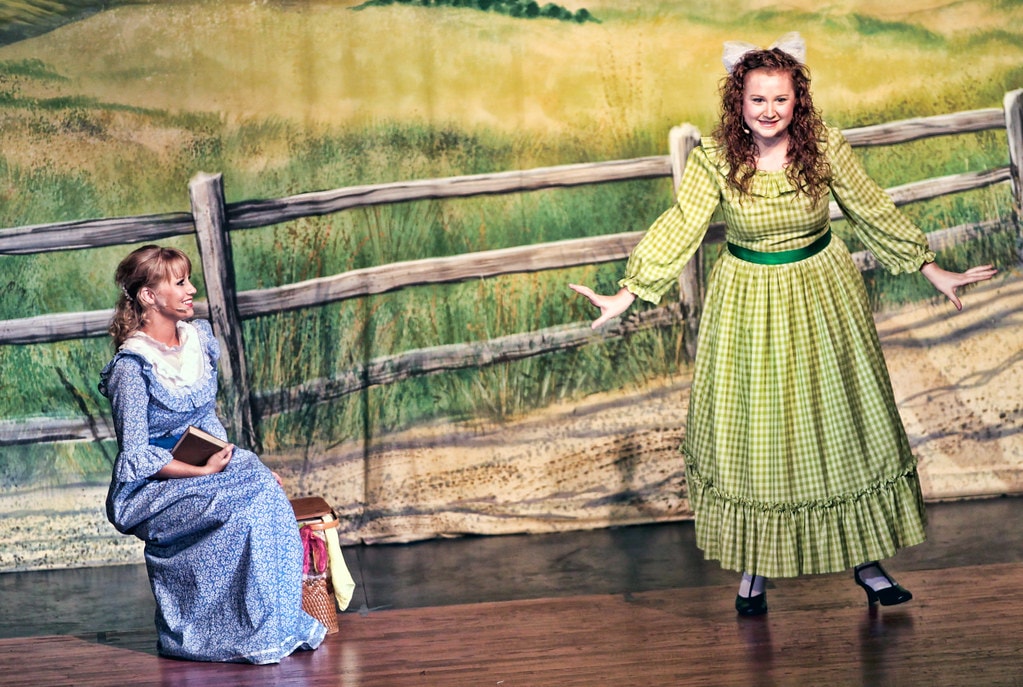 Oklahoma! — Laurey in her blue calico dress sits on a fence rail holding a book and a picnic basket as Ado Annie in her green gingham dress gestures expressively toward her before the painted prairie fence backdrop