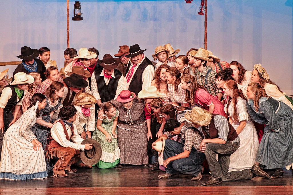 Oklahoma! — the full company finale: over 40 cast members in period frontier costumes crowd the warmly lit stage around Aunt Eller in her pink hat at center, cowboys kneeling in front and the full ensemble of men and women packed together in a joyful celebration scene