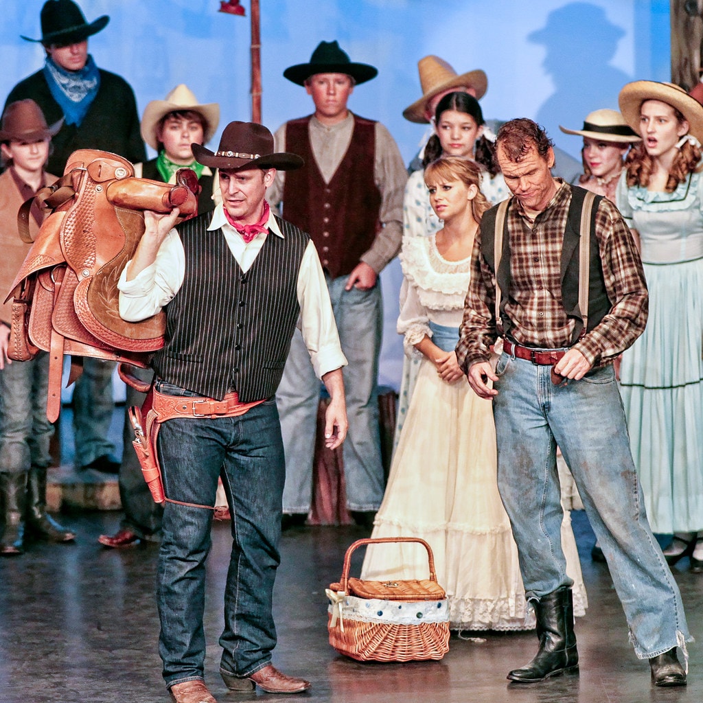 Oklahoma! — the box social confrontation: Curly in his black vest and pink bandana carries a leather saddle over his shoulder as he faces Jud Fry in his plaid shirt and suspenders before the full ensemble of cowboys and women in prairie dresses watching from behind, Laurey in her white ruffled dress and a picnic basket visible between them against the pale blue sky backdrop
