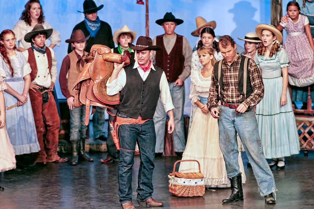 Oklahoma! — the same box social confrontation from a wider angle: Curly with the saddle on his shoulder faces Jud Fry at center stage as the full ensemble of over 20 cast members in period costumes crowds the stage behind them, Laurey in white at center and the picnic basket on the floor between the two men