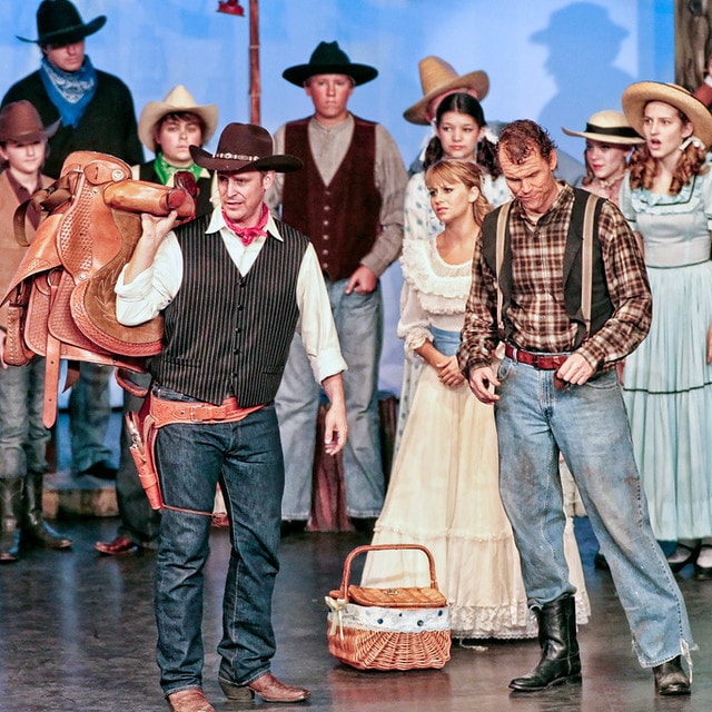 Oklahoma! — a tighter crop of the box social scene: Curly in his black vest and pink bandana holds the saddle at left as Jud Fry in his plaid shirt and suspenders leans forward at right, Laurey in her white ruffled dress and the wicker picnic basket visible between them, the ensemble watching from behind