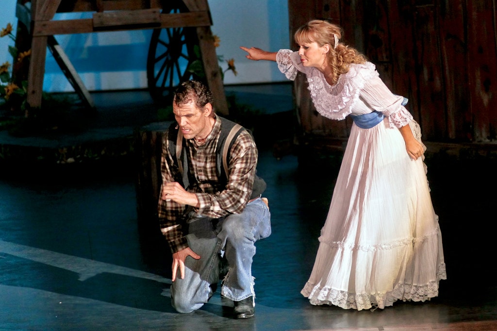Oklahoma! — Laurey confronts Jud Fry: Laurey in her white lace-trimmed dress leans forward with one arm outstretched pointing accusingly at Jud Fry who crouches low on the bare stage, the barn set with a wagon wheel and sunflowers visible in the background