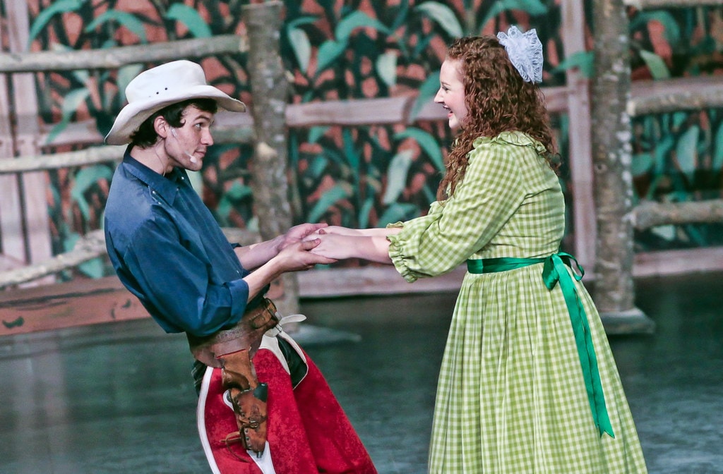 Oklahoma! — Curly in his white cowboy hat and red chaps holds both of Ado Annie's hands as they face each other smiling before the cornfield fence backdrop