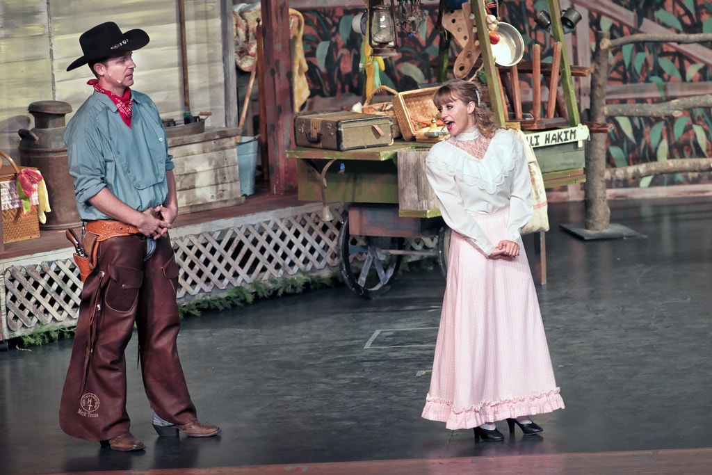 Oklahoma! — Will Parker in his black cowboy hat and brown leather chaps stands beside Ado Annie in her pink ruffled dress as she reacts with delight before the Ali Hakim peddler wagon on the farmyard set