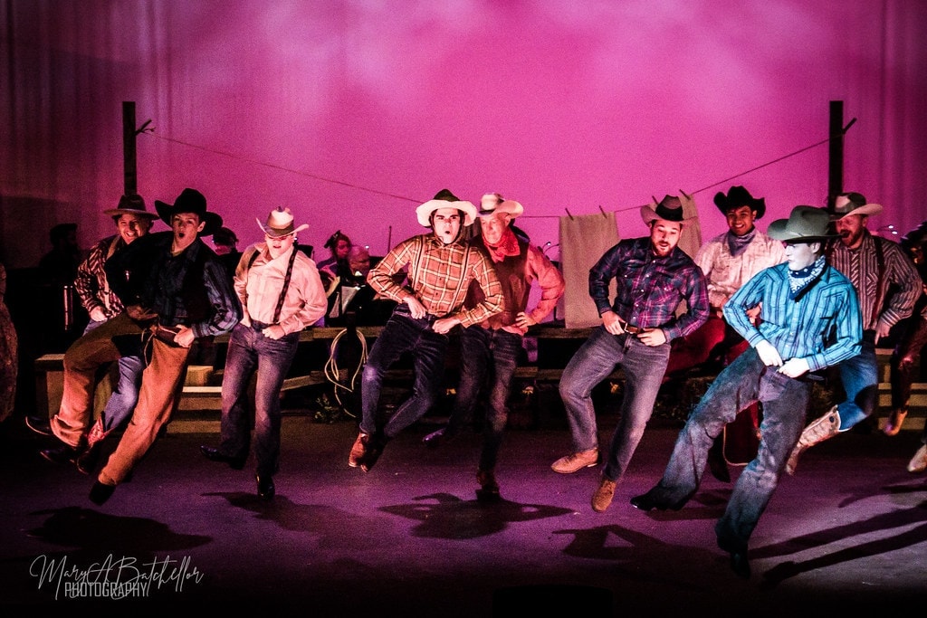 Oklahoma! — cowboys raising hats in celebration under pink lights