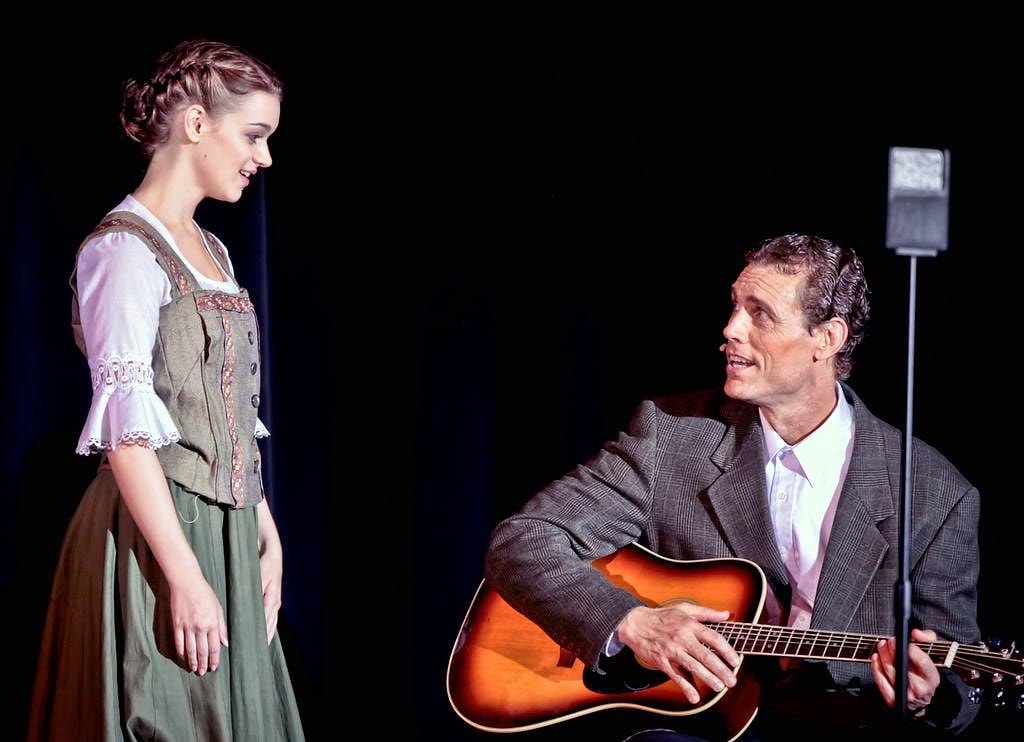 The Sound of Music — Maria in a grey dirndl vest and white blouse faces Captain von Trapp seated with a guitar against a black backdrop in an intimate two-person scene