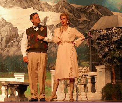 The Sound of Music — a man in an argyle vest gestures toward a woman in a pink dress who stands with one hand on her hip before the painted alpine mountain backdrop