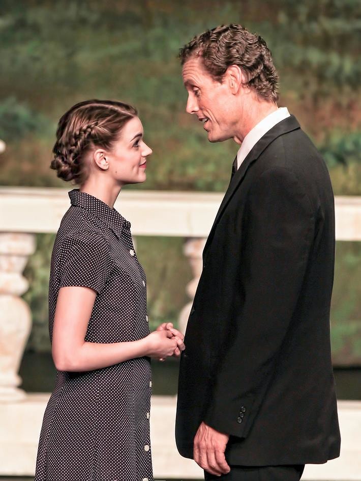 The Sound of Music — Maria in a black polka-dot dress holds hands with Captain von Trapp in a black suit before the painted garden terrace backdrop in a tender romantic scene