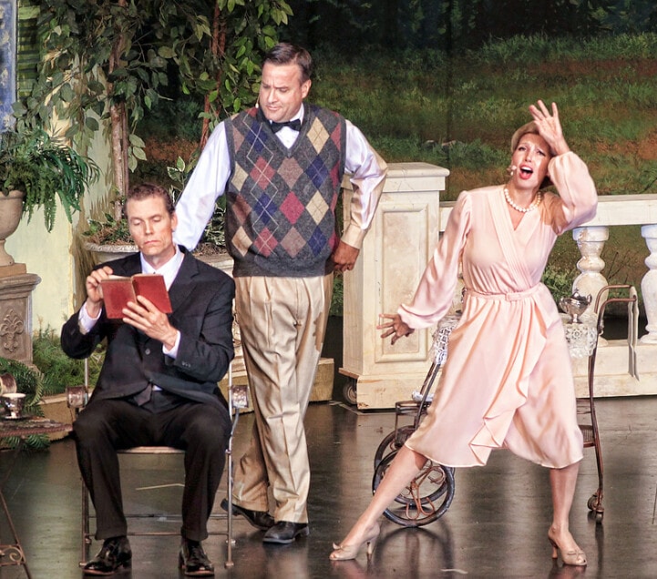 The Sound of Music — three performers on the garden terrace set: a man in a black suit reads a book, a man in an argyle vest stands behind him, and a woman in a pink dress sings dramatically with one arm raised