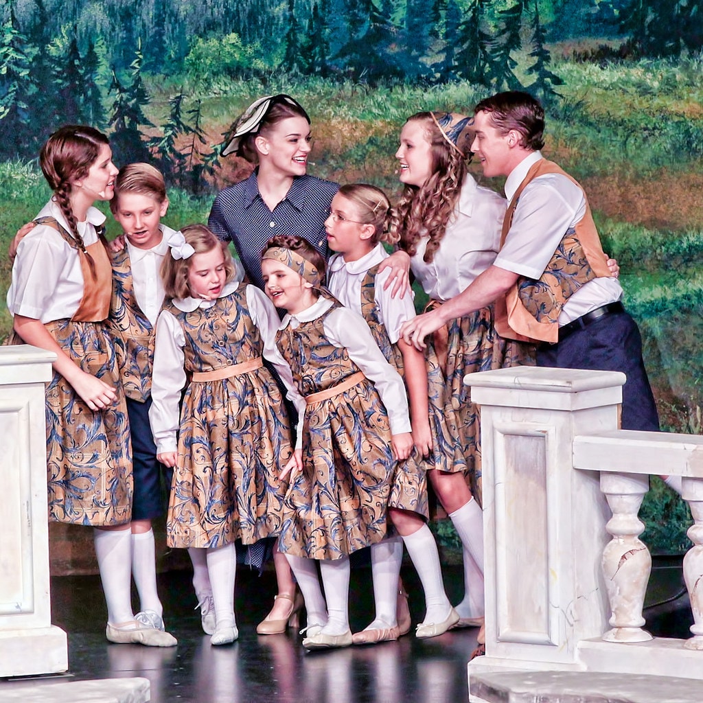 The Sound of Music — Maria in a blue polka-dot dress and the older von Trapp son gather the seven children in matching gold-and-blue patterned outfits before the painted alpine forest backdrop