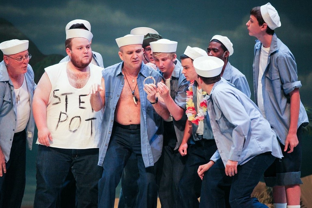 South Pacific — a shirtless man with a necklace holds up a ring as a group of sailors in white caps and blue shirts crowd around him curiously against a stormy tropical backdrop