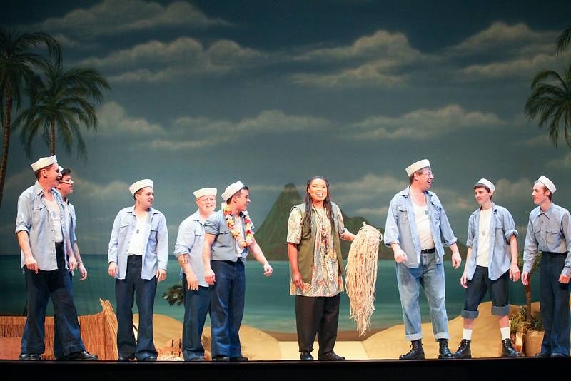 South Pacific — a woman in a floral shirt holding a mop stands center stage surrounded by sailors in white caps and blue shirts laughing together before a painted tropical beach backdrop with palm trees