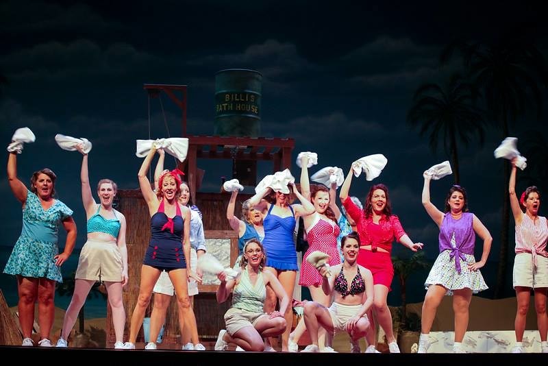 South Pacific — the full female ensemble in colorful 1940s beach costumes raises white towels joyfully before the Billis Bath House set against a dark stormy sky backdrop
