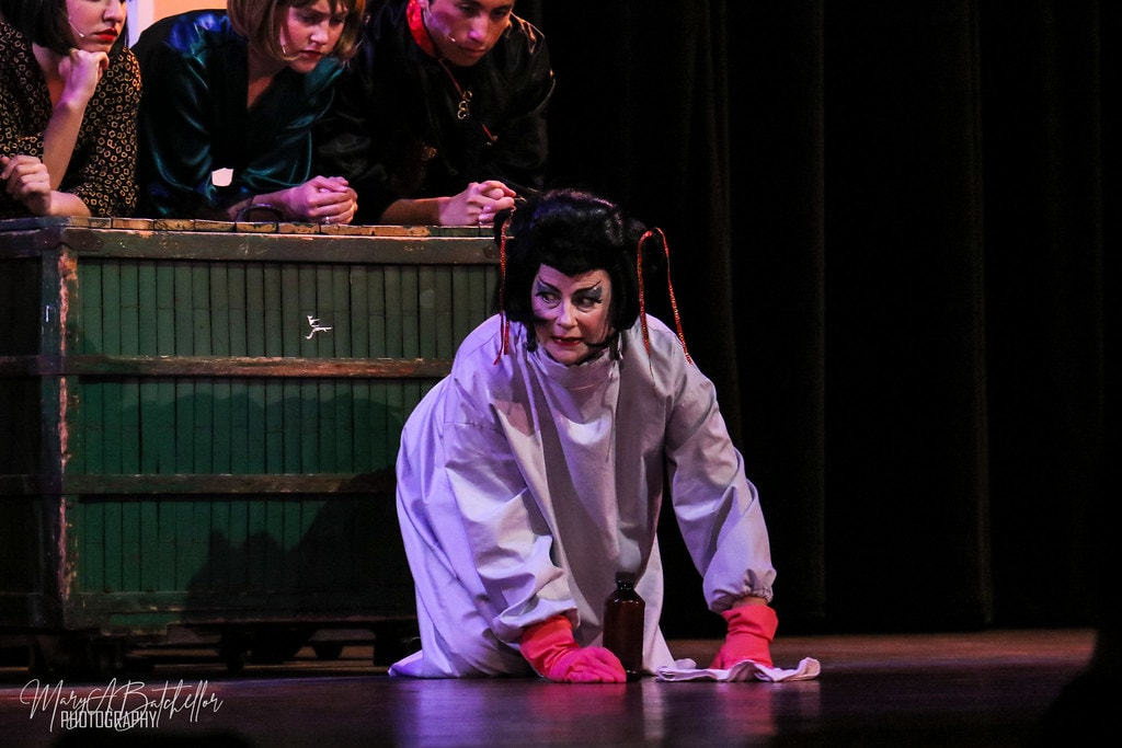 Thoroughly Modern Millie — Mrs. Meers in a white costume crouches on the stage floor as hotel guests look on