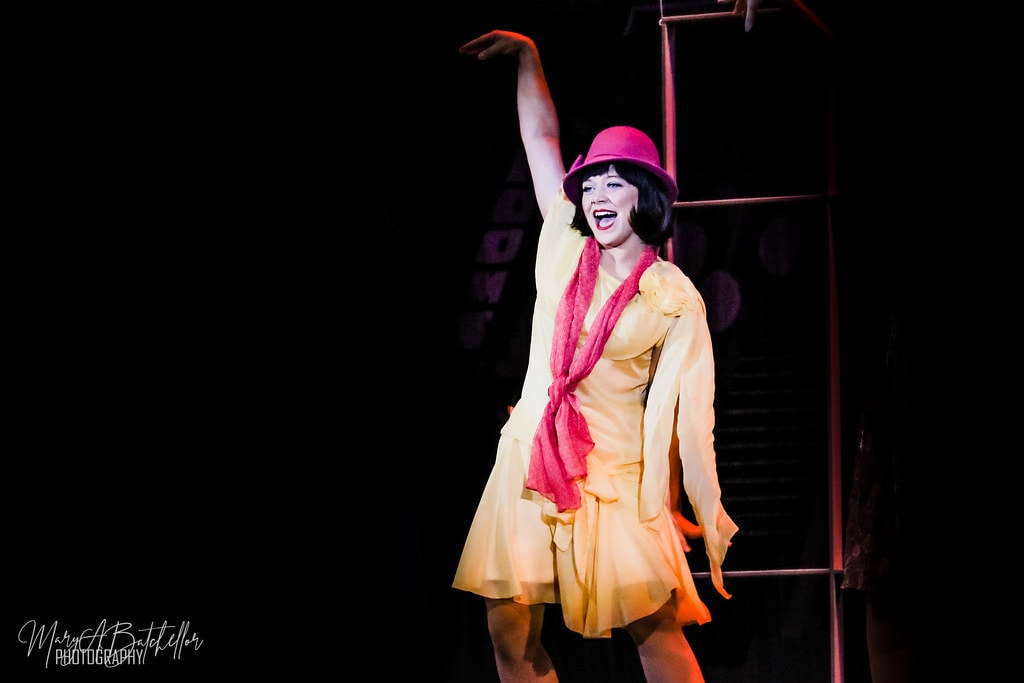 Thoroughly Modern Millie — Millie in a yellow dress and pink bowler hat belts a solo with one arm raised