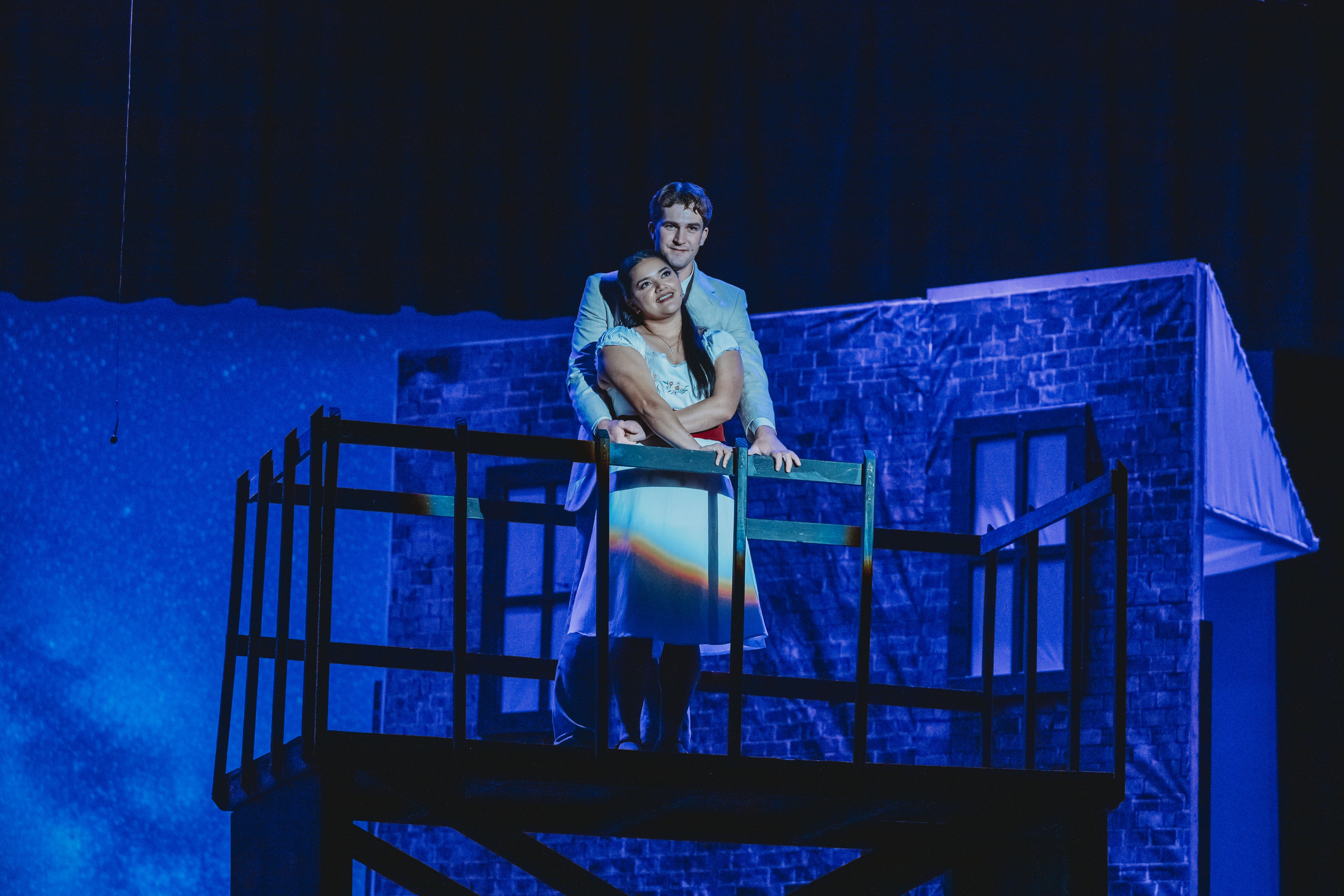 West Side Story 2026 - Maria and Tony embracing on the fire escape balcony with blue starlit backdrop