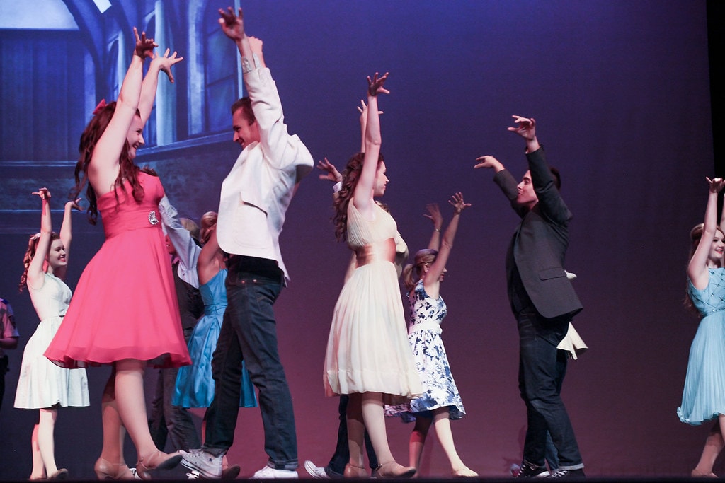 Full ensemble with arms raised blue arch backdrop — West Side Story